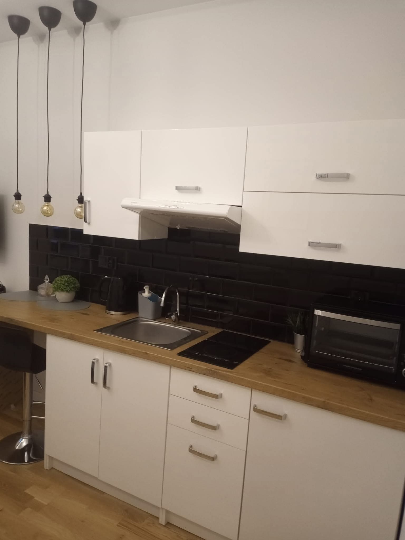 Biała zabudowa kuchenna z drewnianym blatem i czarnymi płytkami na ścianie. Nad blatem wiszą trzy lampy z żarówkami. Widoczny zlew, płyta grzewcza i sprzęt AGD.