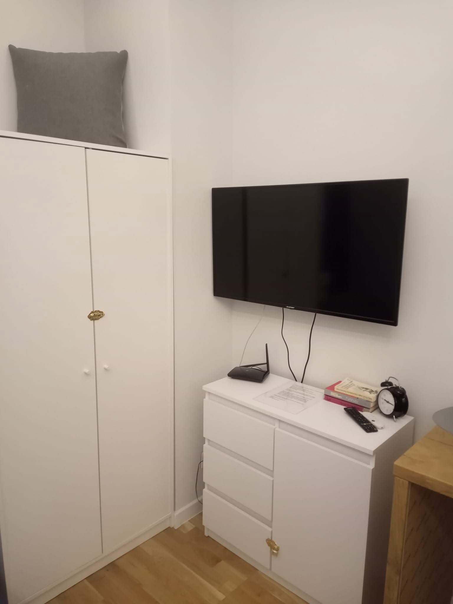 Biała szafa z szarą poduszką na górze, obok komoda z telewizorem na ścianie, routerem, książkami i budzikiem. Minimalistyczne wnętrze pokoju.