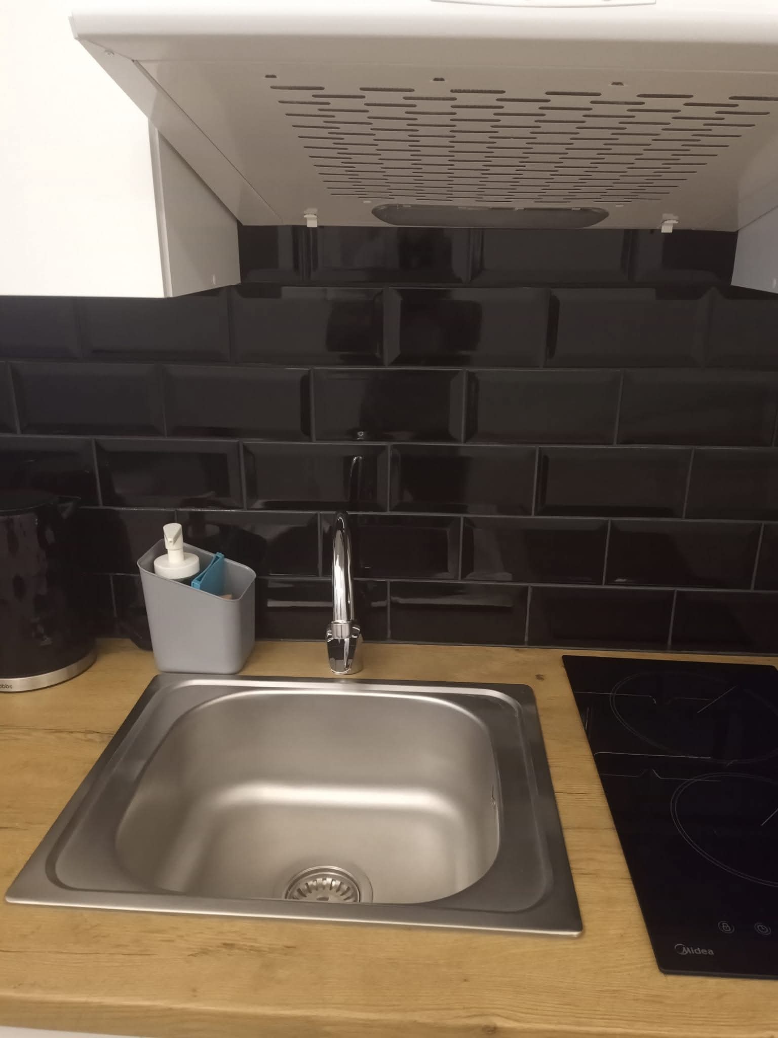 Stalowy zlew wkomponowany w drewniany blat, bateria, czarne, błyszczące płytki na ścianie i okap kuchenny. Nowoczesna aranżacja kuchni.