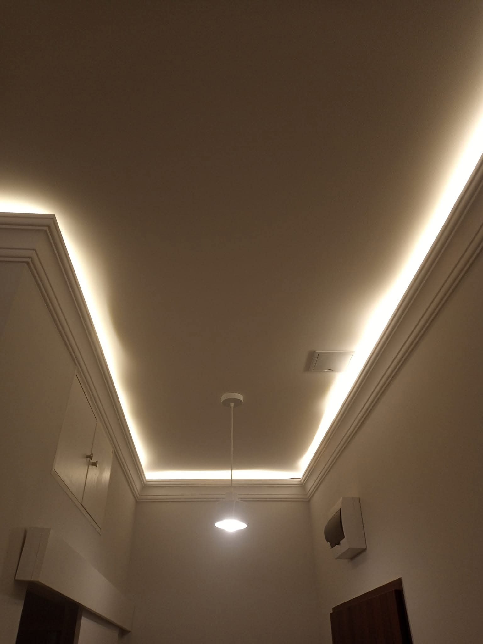 Nowoczesne oświetlenie sufitowe LED w kremowym wnętrzu. Dekoracyjne listwy i centralna lampa wisząca tworzą unikalny klimat. Widok z dołu.