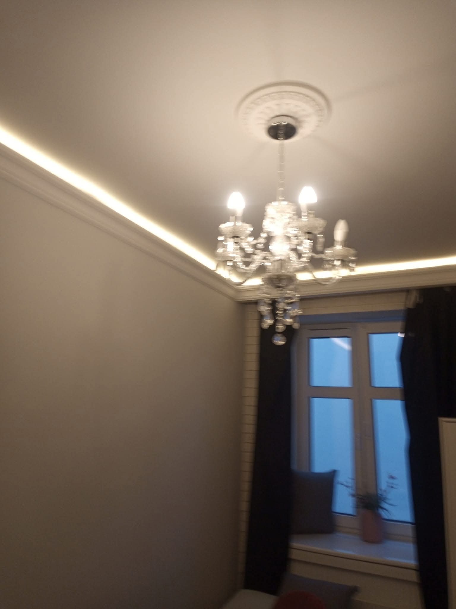 Kryształowy żyrandol pod sufitem z dekoracyjną rozetą i listwami LED. Okno z zasłonami w tle. Subtelne oświetlenie wnętrza.