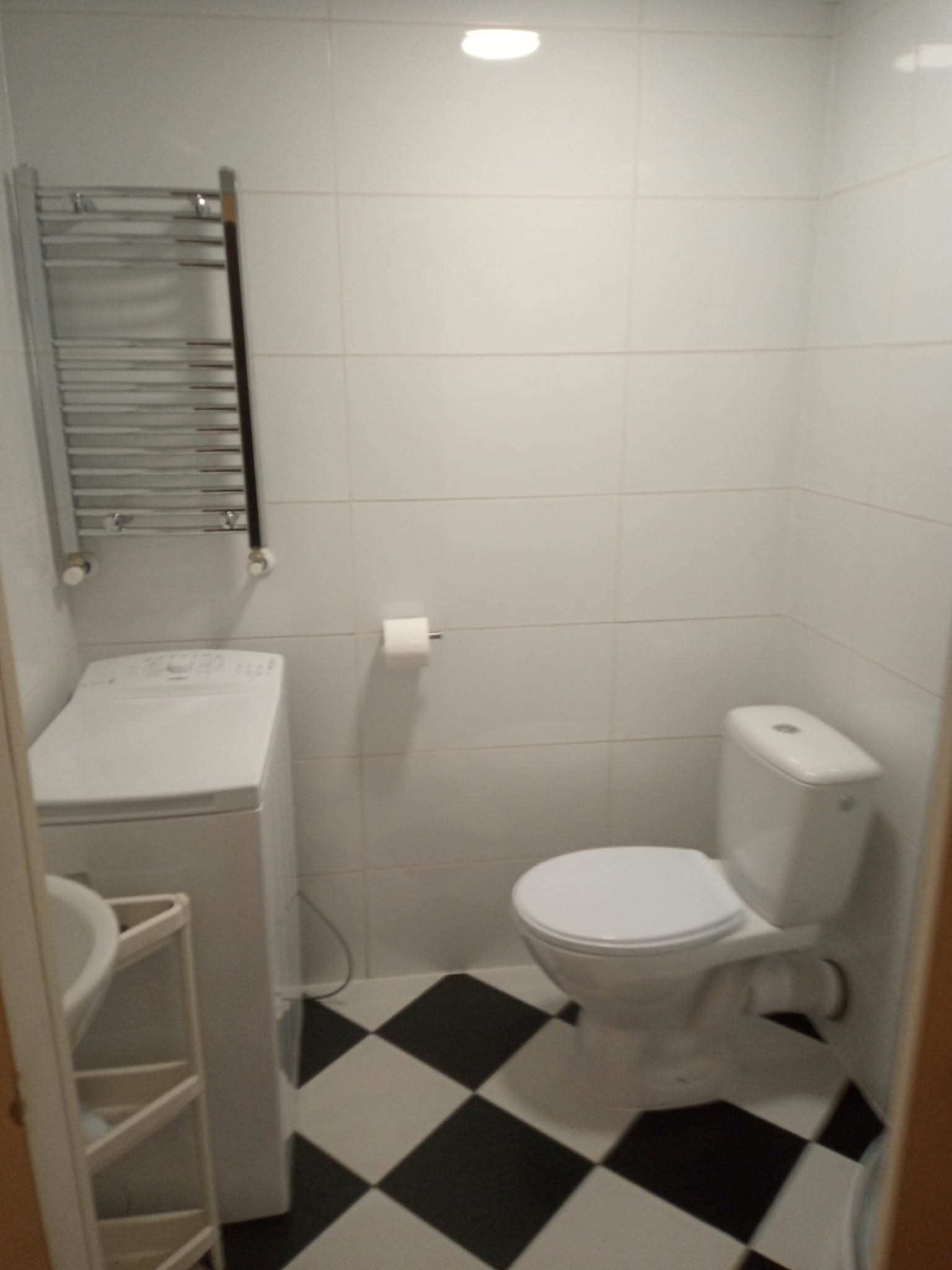 Biała łazienka z czarno-białą szachownicą na podłodze. Widoczna toaleta, pralka, grzejnik drabinkowy i umywalka. Minimalistyczny wystrój, jasne oświetlenie.