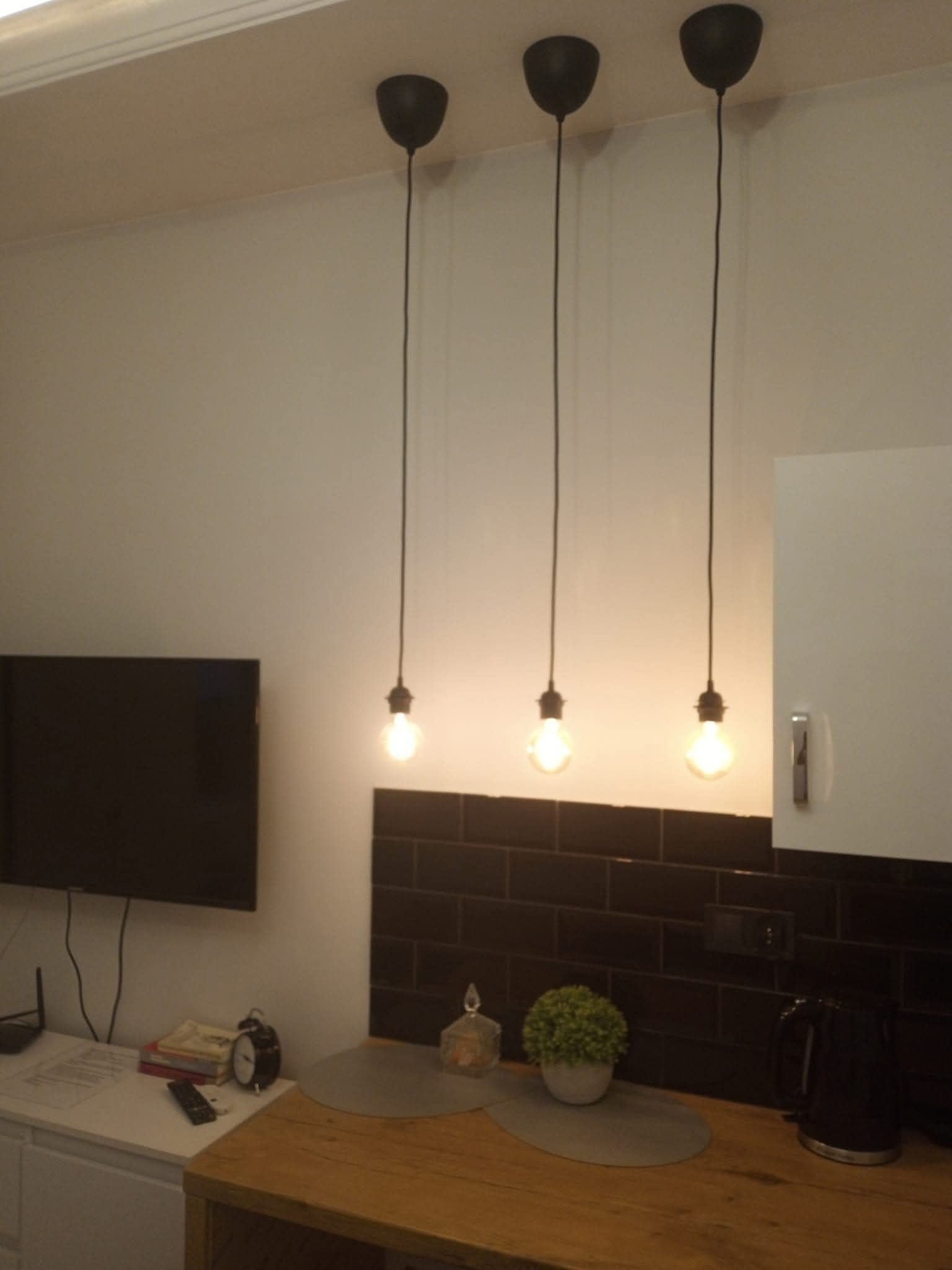 Nowoczesna kuchnia z trzema wiszącymi lampami z żarówkami Edisona, czarne płytki na ścianie, drewniany blat i białe szafki. Minimalistyczny design, ciepłe światło.