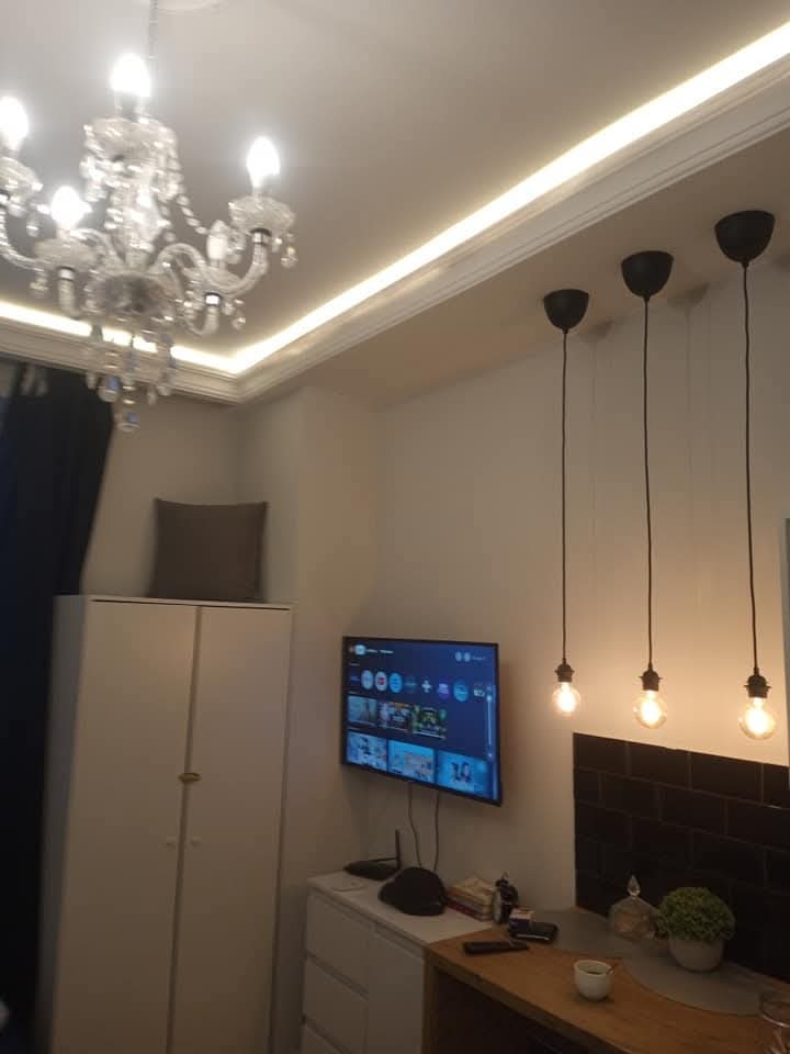 Nowoczesne oświetlenie wnętrza: kryształowy żyrandol, listwa LED podsufitowa i trzy wiszące lampy z żarówkami Edisona. Telewizor na ścianie, białe meble.