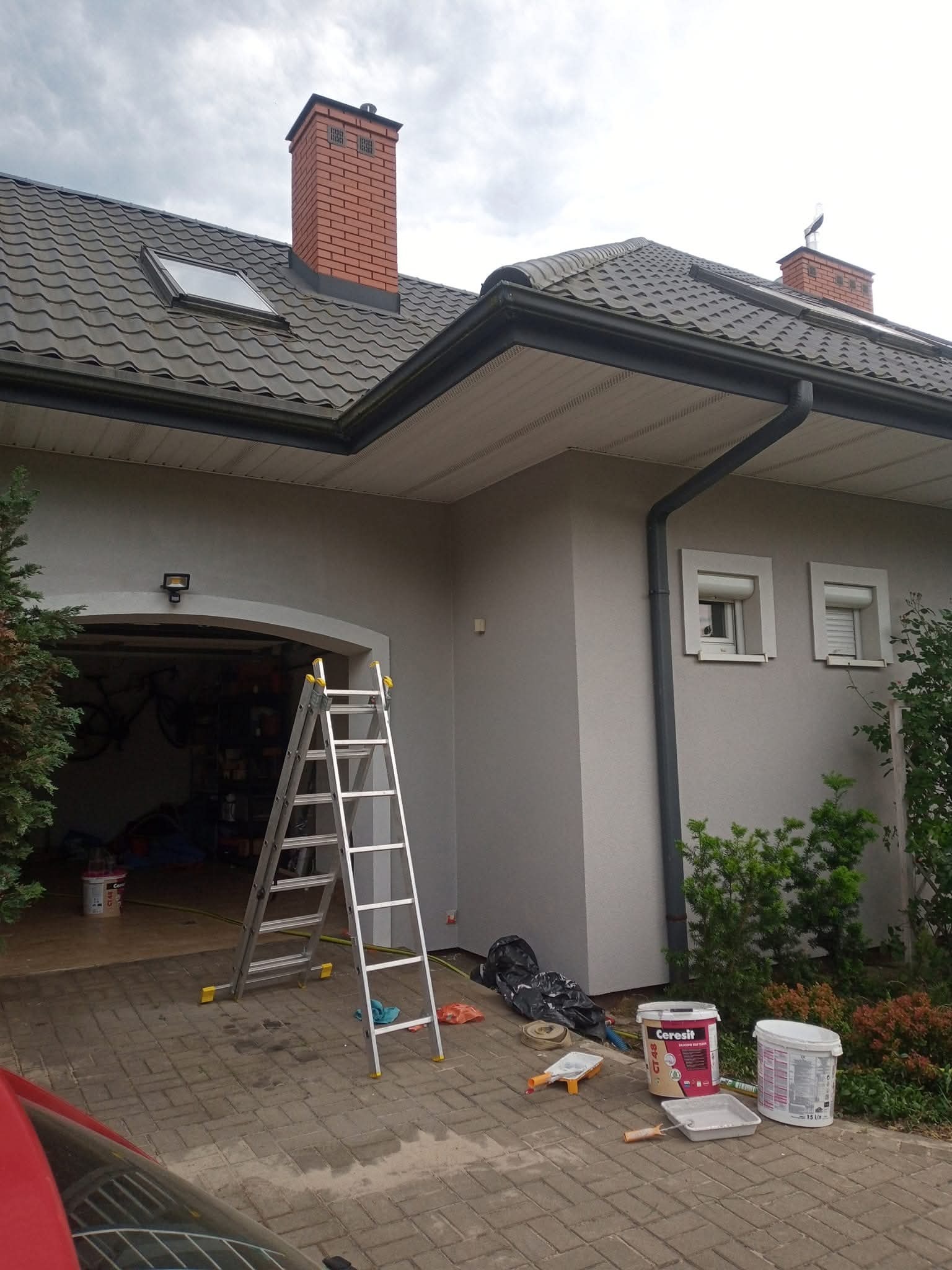 Dom jednorodzinny po malowaniu elewacji, widoczna drabina, pędzle i farby. Świeżo pomalowana ściana w odcieniach szarości. Dachówka ceramiczna i ceglany komin.