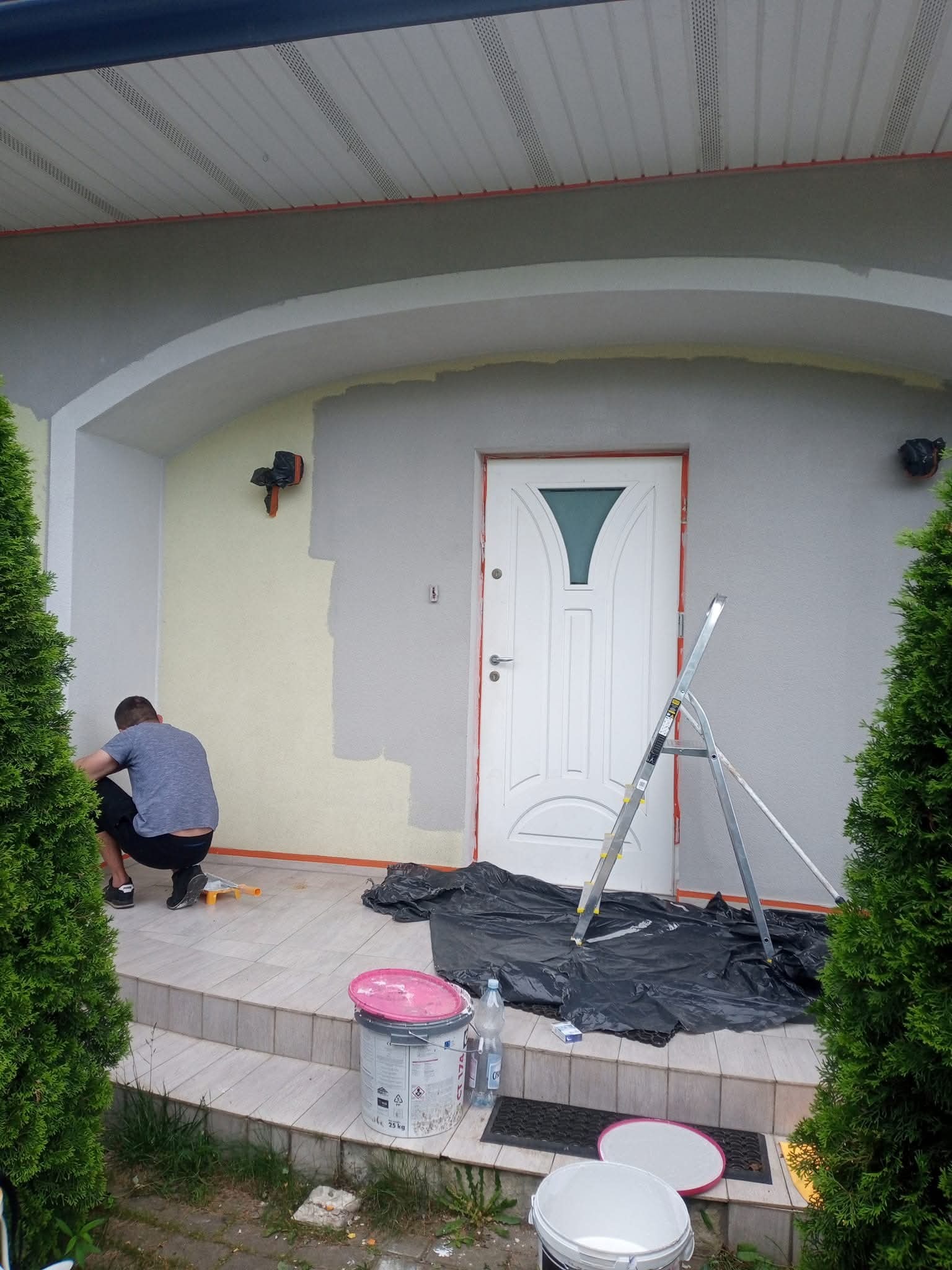 Malowanie elewacji domu: mężczyzna w trakcie pracy, widoczne pędzle, drabina, farba i zabezpieczona folią strefa malowania. Częściowo pomalowana ściana.