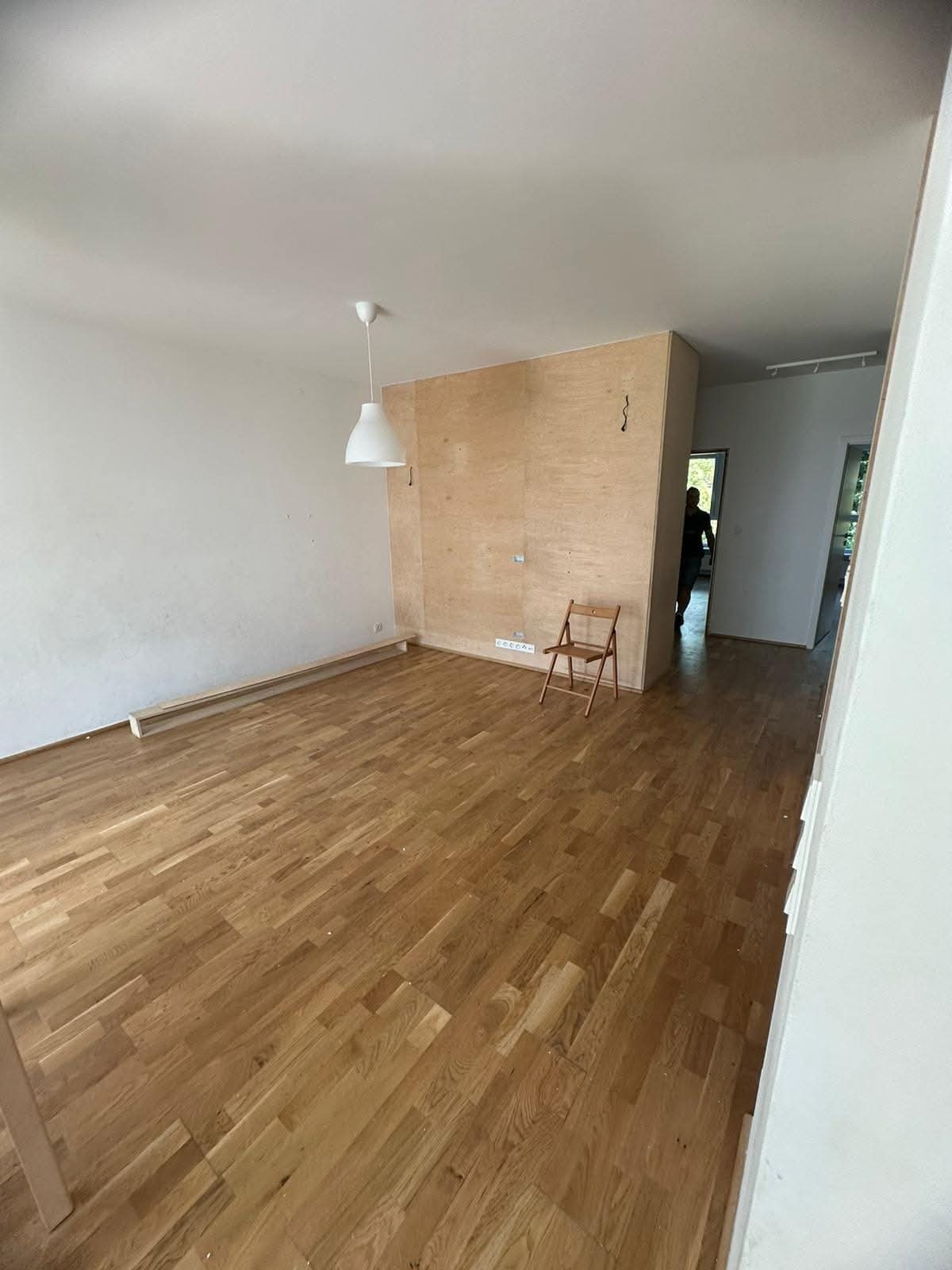 Wnętrze pokoju z drewnianą podłogą i ścianą z płyty. Widoczny fragment listwy przypodłogowej, krzesło i lampa wisząca. W tle osoba w przejściu do innego pomieszczenia.