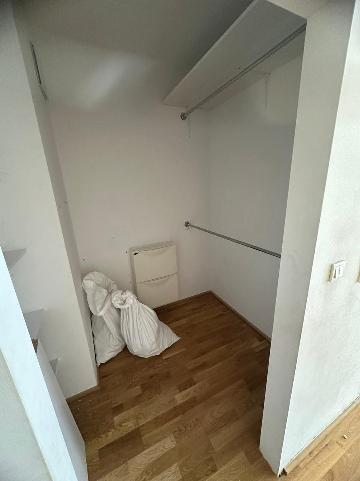 Wnęka garderoby z drewnianą podłogą i półkami. Dwa worki budowlane stoją w rogu. Widoczna szafka na buty i karnisz na ubrania. Świeżo wykończone wnętrze.