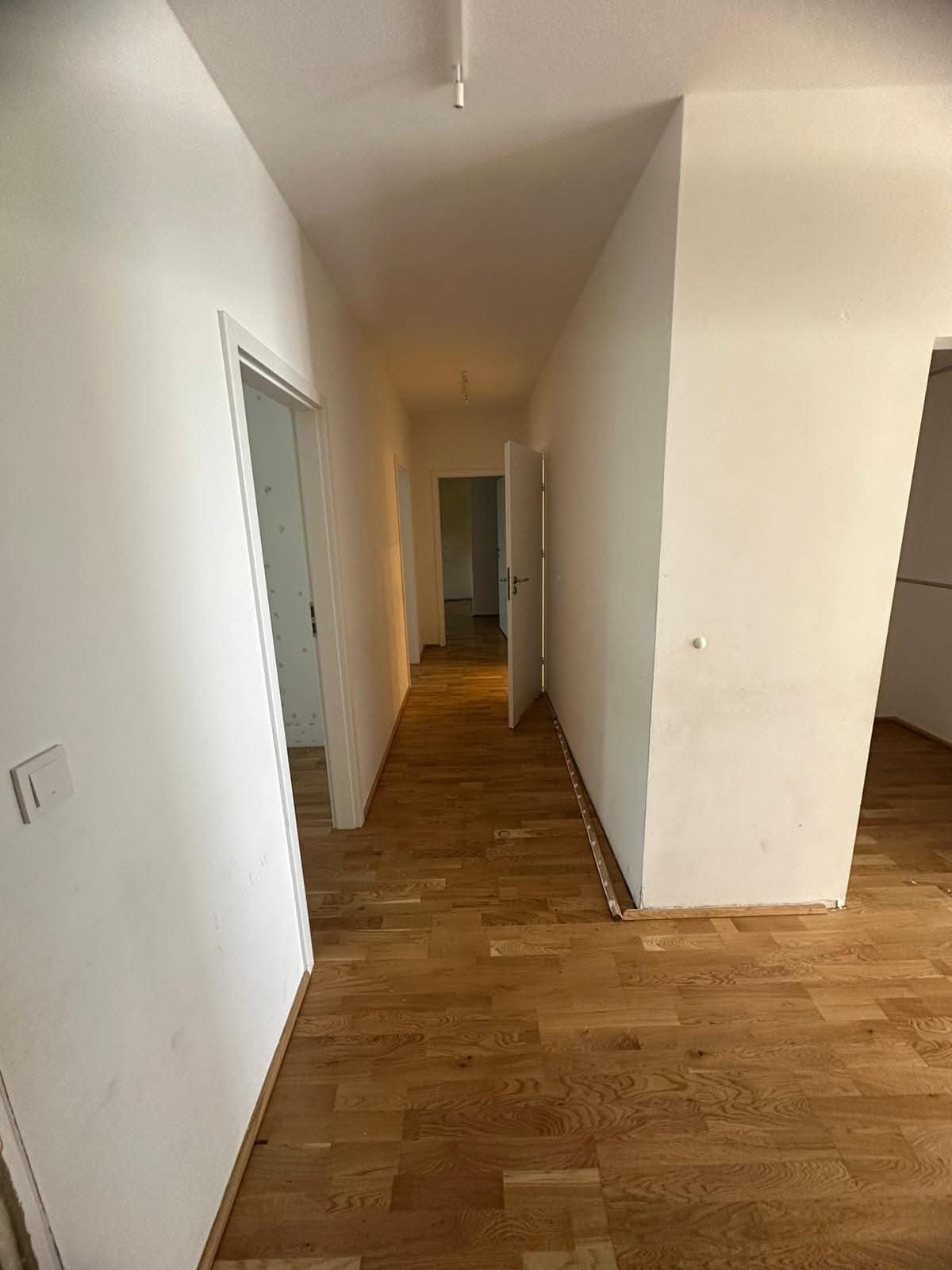 Korytarz z drewnianą podłogą i białymi ścianami. Widoczne otwarte drzwi i fragmenty listew przypodłogowych. Perspektywa wnętrza z widokiem na kolejne pomieszczenia.