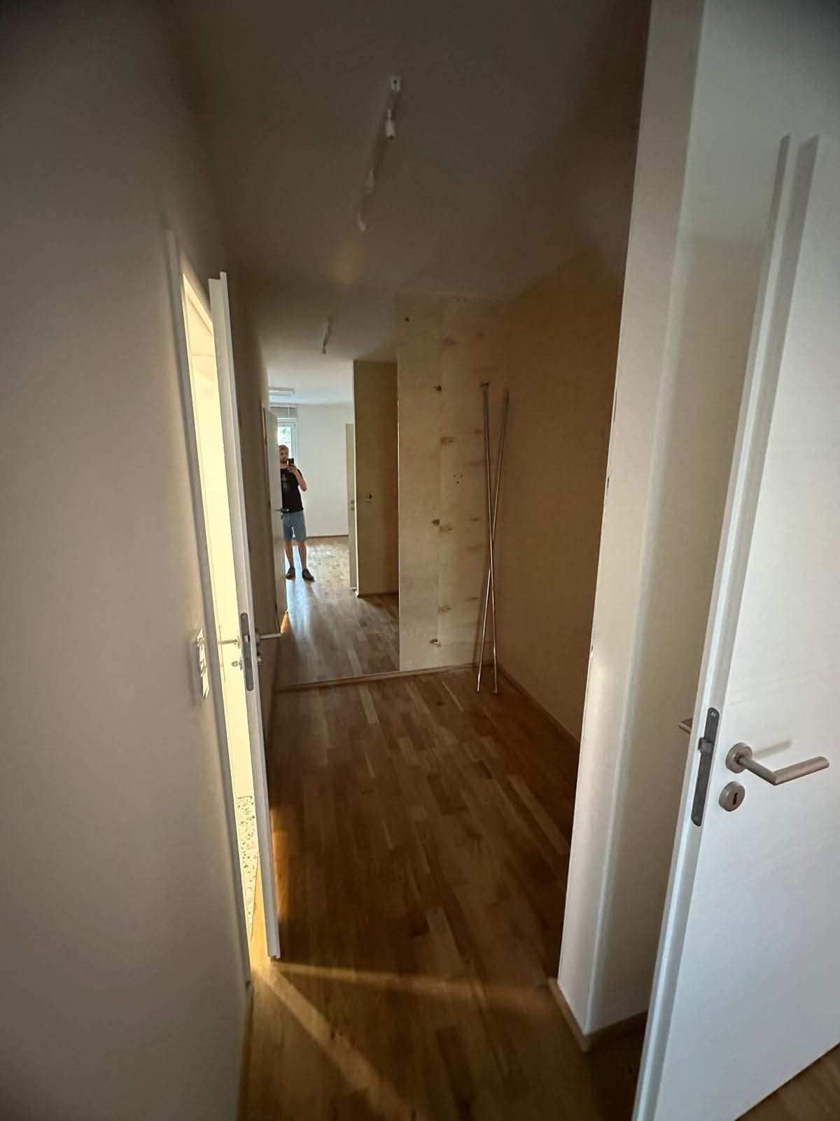 Korytarz po remoncie z drewnianą podłogą i białymi drzwiami. Widoczne ślady po demontażu ściany działowej. W lustrze odbija się osoba robiąca zdjęcie.