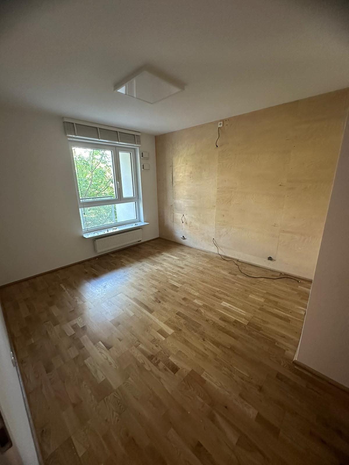 Pokój przed remontem: parkiet, ściany przygotowane do malowania, widoczne kable. Okno z widokiem na zieleń. Minimalistyczna lampa sufitowa. Jasne wnętrze.
