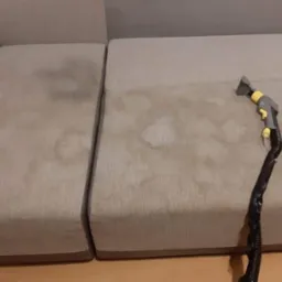 Beżowa sofa z widocznymi plamami czyszczona parownicą z szarą rączką i żółtymi detalami, ukazująca proces usuwania zabrudzeń.