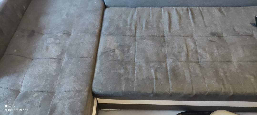 Fragment szarej, narożnej sofy tapicerowanej z widocznymi plamami i zabrudzeniami na siedzisku i oparciu.
