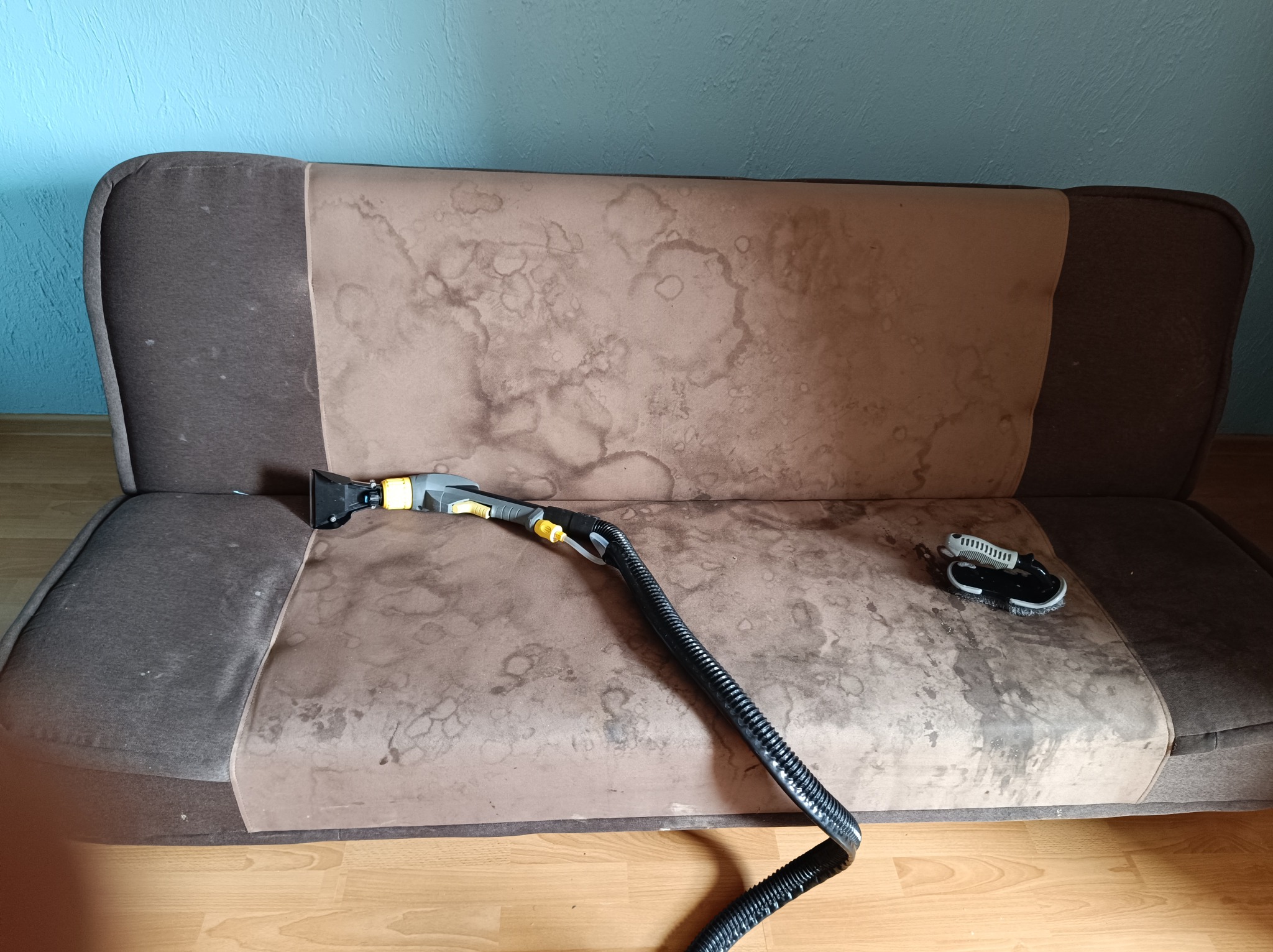 Brudna, dwukolorowa kanapa z widocznymi plamami, częściowo wyczyszczona za pomocą odkurzacza piorącego z czarnym wężem i małej szczotki.