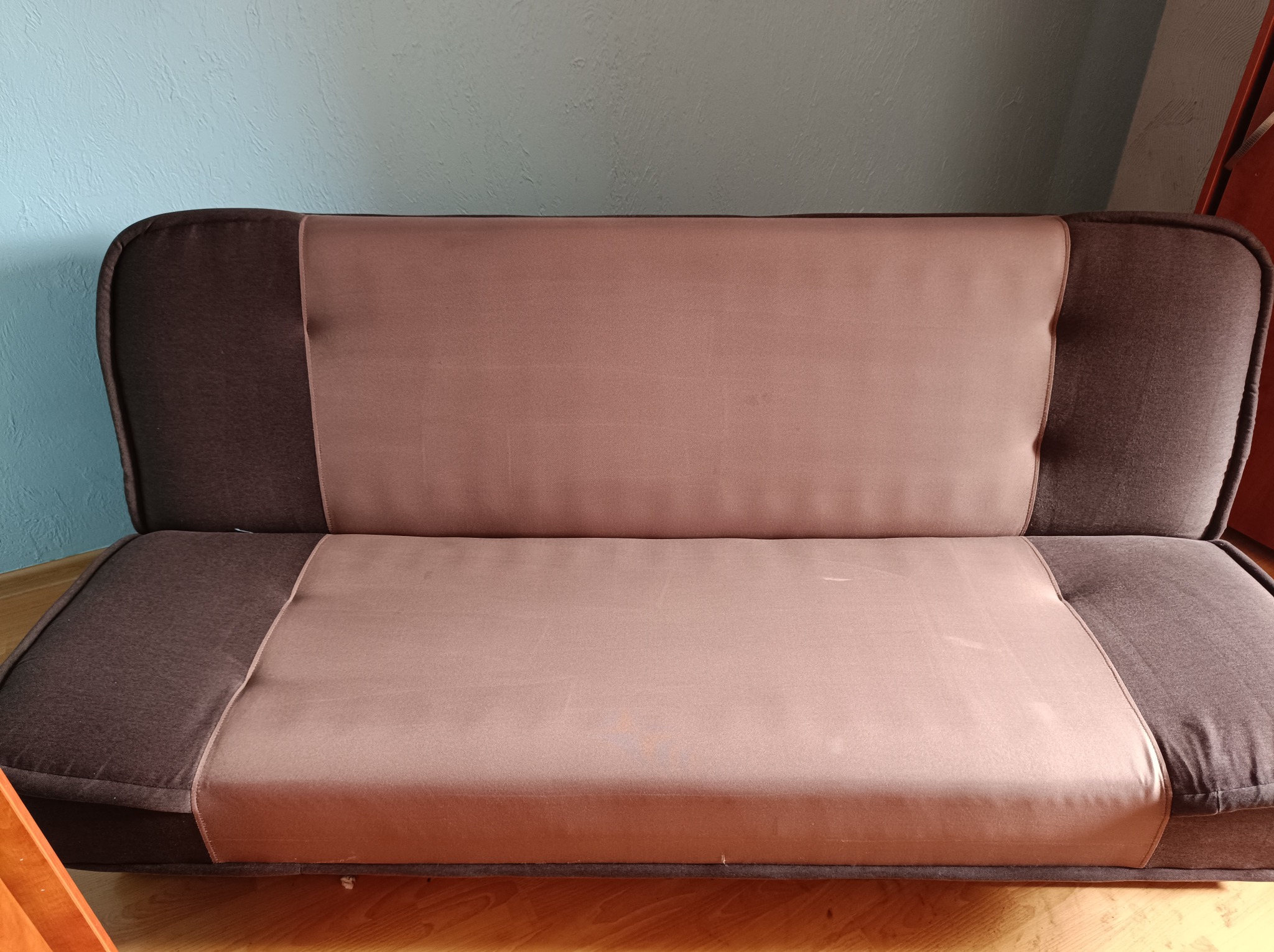 Rozłożona sofa z brązowymi bokami i różowym siedziskiem na drewnianej podłodze, tło stanowi niebieska ściana.