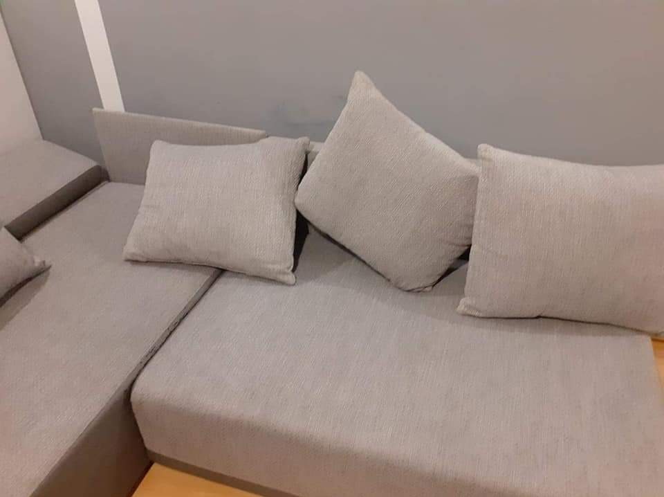 Jasnoszara sofa narożna z poduszkami o wyraźnej fakturze materiału, ustawiona pod szarą ścianą.