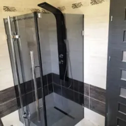 Nowoczesna kabina prysznicowa z czarnym panelem natryskowym i szklanymi drzwiami, w łazience wykończonej płytkami w odcieniach beżu i brązu z dekoracyjnym pasem.