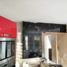 Częściowo wyłożona ściana czarnym marmurem z białymi żyłami w kuchni. Widoczne krzyżyki dystansowe, szafka i sprzęt AGD. Etap prac wykończeniowych.