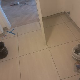 Paweł Lelek - Ułożone beżowe płytki podłogowe w przedpokoju, widoczne narzędzia i fragment podłogi w innym wzorze. Praca w toku, detale wykończeniowe.