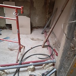 Remont w toku: widok na podłogę z rurami, przewodami, pianką montażową i rusztowaniem. Ściany w stanie surowym, płyty gipsowe częściowo zamontowane. Chaos remontowy.