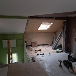 Wnętrze w trakcie przebudowy: widoczne odsłonięte ściany, okno dachowe, narzędzia i materiały budowlane. Fragment zielonej ściany z grzejnikiem.