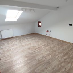 Paweł Lelek - Jasne panele podłogowe w pokoju na poddaszu z oknem dachowym i białymi ścianami. Widoczne grzejnik i instalacja. Uwagę zwraca skośny sufit i oświetlenie.