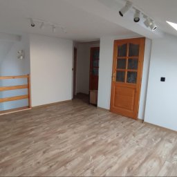 Paweł Lelek - Jasne poddasze po remoncie: świeżo ułożone panele podłogowe w odcieniach drewna, białe ściany z oknem dachowym i drewniane drzwi z przeszkleniami.