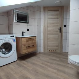 Paweł Lelek - Jasna łazienka na poddaszu z pralką, drewnianą szafką pod umywalką i podświetlanym lustrem. Ukończone płytki na ścianach i drewnopodobna podłoga.