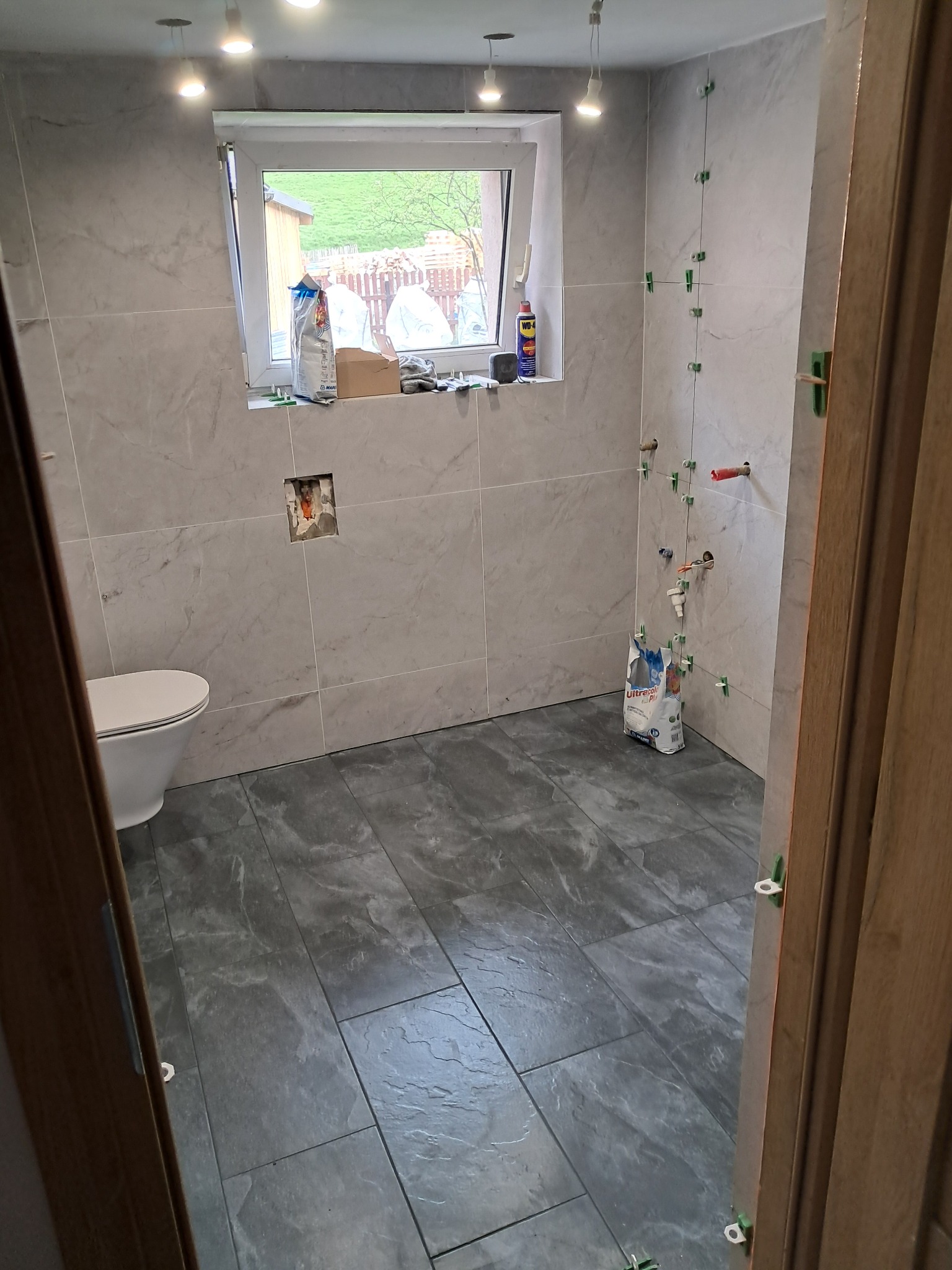 Łazienka w trakcie wykańczania: szare płytki na podłodze i ścianach, widoczne akcesoria glazurnicze, okno z widokiem na zewnątrz. Częściowo zamontowana toaleta.