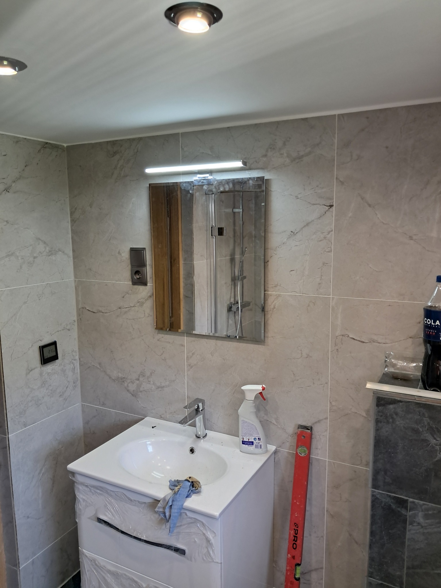 Ukończona łazienka z marmurowymi płytkami, białą szafką umywalkową i lustrem z oświetleniem. Na blacie leży ściereczka, a obok stoi poziomica.