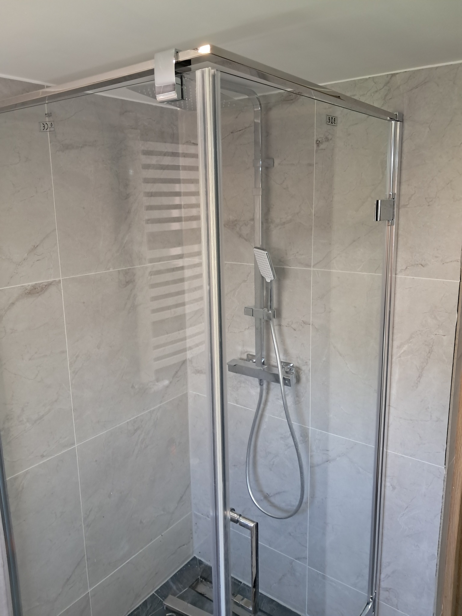 Szklana kabina prysznicowa z chromowanymi detalami na tle ściany wyłożonej szarymi, marmurkowymi płytkami. Widoczna armatura prysznicowa i fragment grzejnika.