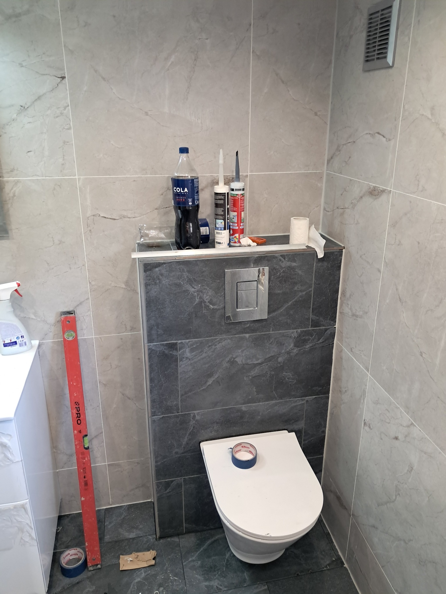 Wykończona toaleta w łazience z szarymi płytkami imitującymi marmur na ścianach i zabudowie, widoczny poziomica, taśma malarska i akcesoria montażowe.