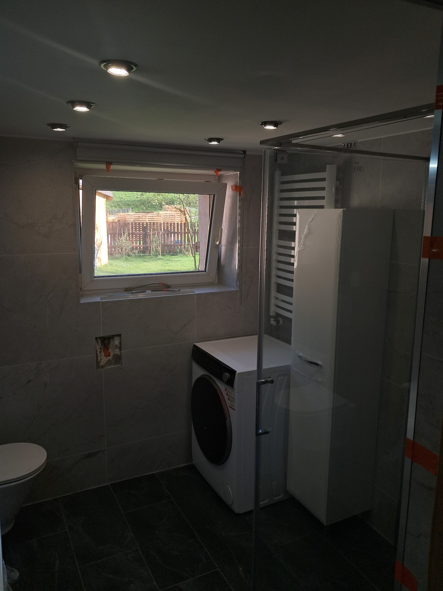 Wykończona łazienka w szarościach: płytki imitujące kamień, kabina prysznicowa ze szklaną ścianką, pralka i okno z widokiem na ogród. Widoczne detale wykończeniowe.