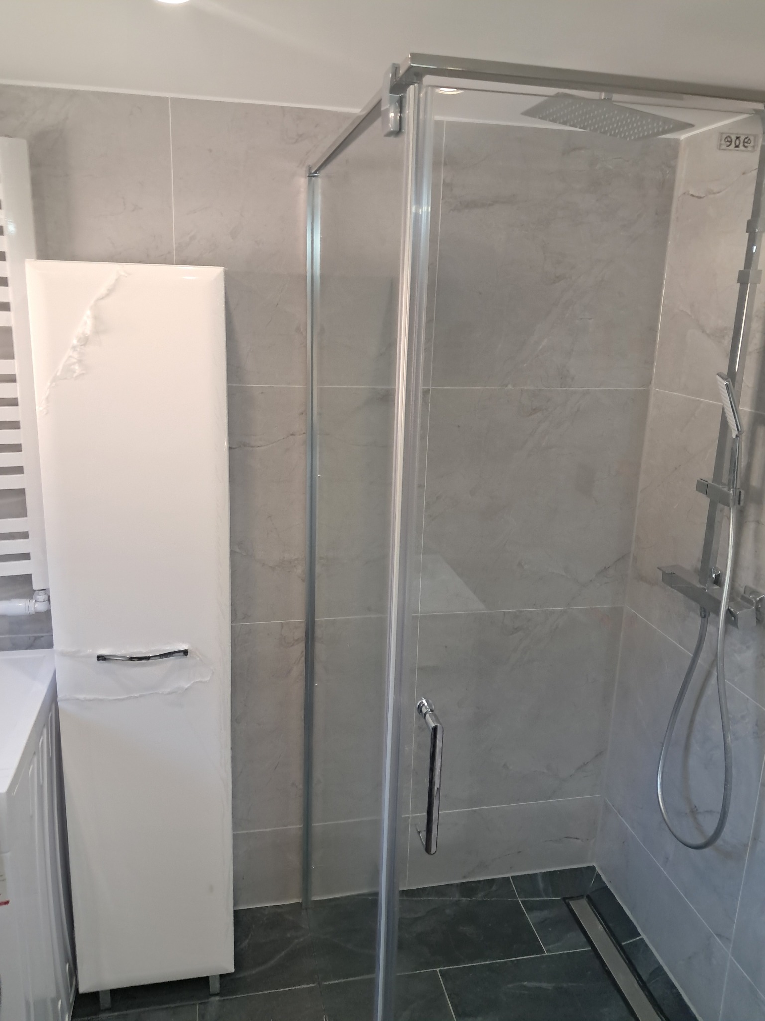 Nowoczesna kabina prysznicowa z szarymi płytkami imitującymi marmur. Biała szafka łazienkowa obok pralki, całość utrzymana w minimalistycznym stylu.