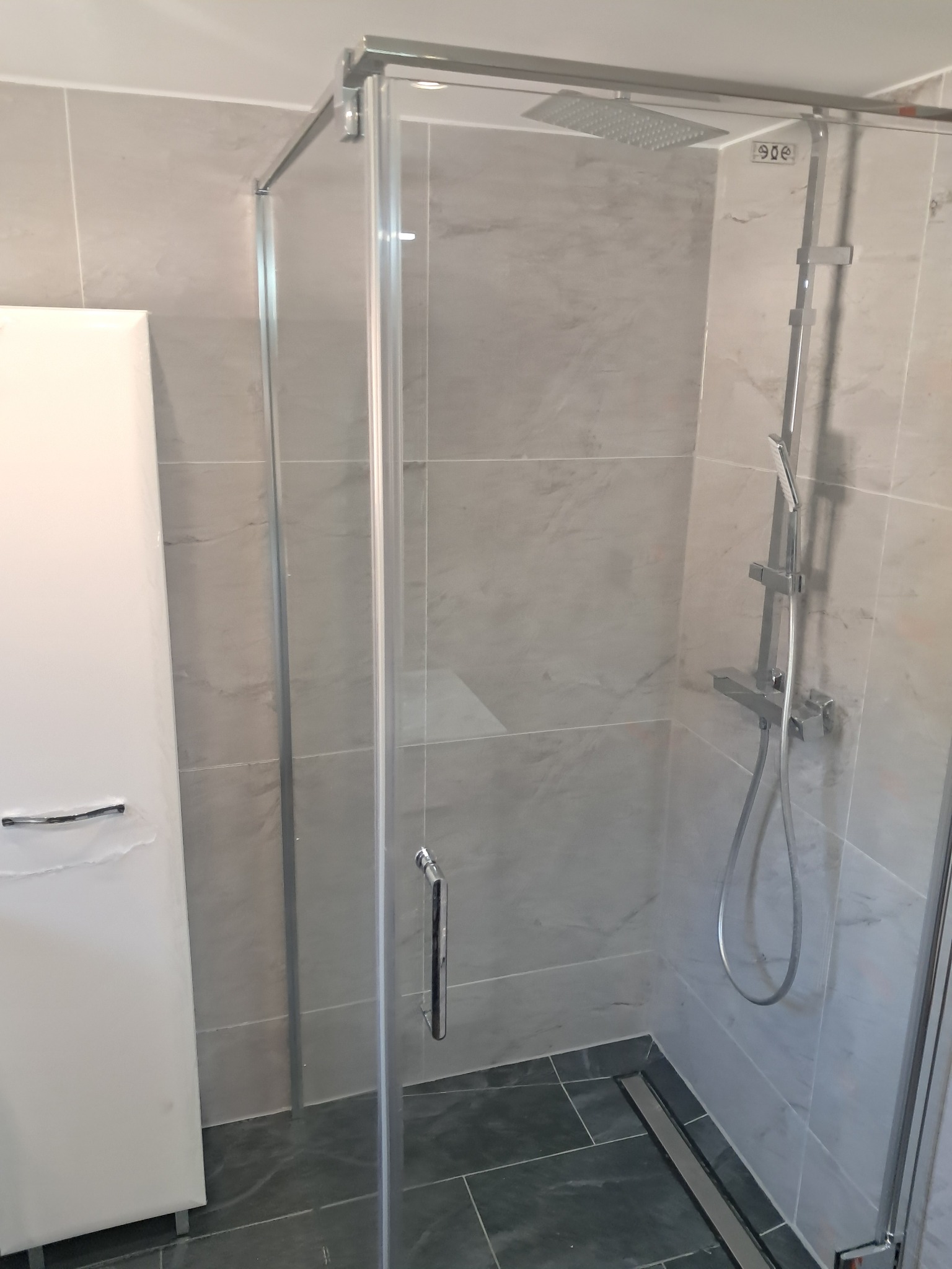 Nowoczesna kabina prysznicowa z deszczownicą i odpływem liniowym, wykończona szarymi płytkami imitującymi marmur i białą szafką.