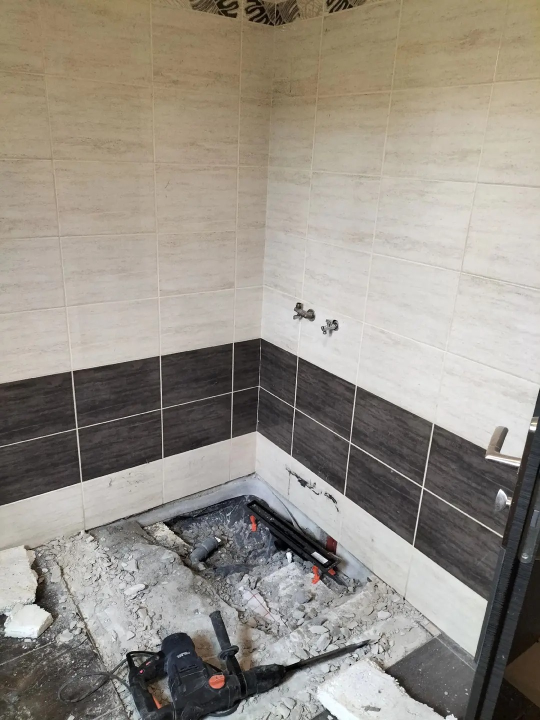 Demontaż podłogi i części płytek w łazience, widoczne rury, gruz i młot udarowy. Etap przygotowawczy do remontu, z widocznymi zaworami i częściowo wykończonymi ścianami.