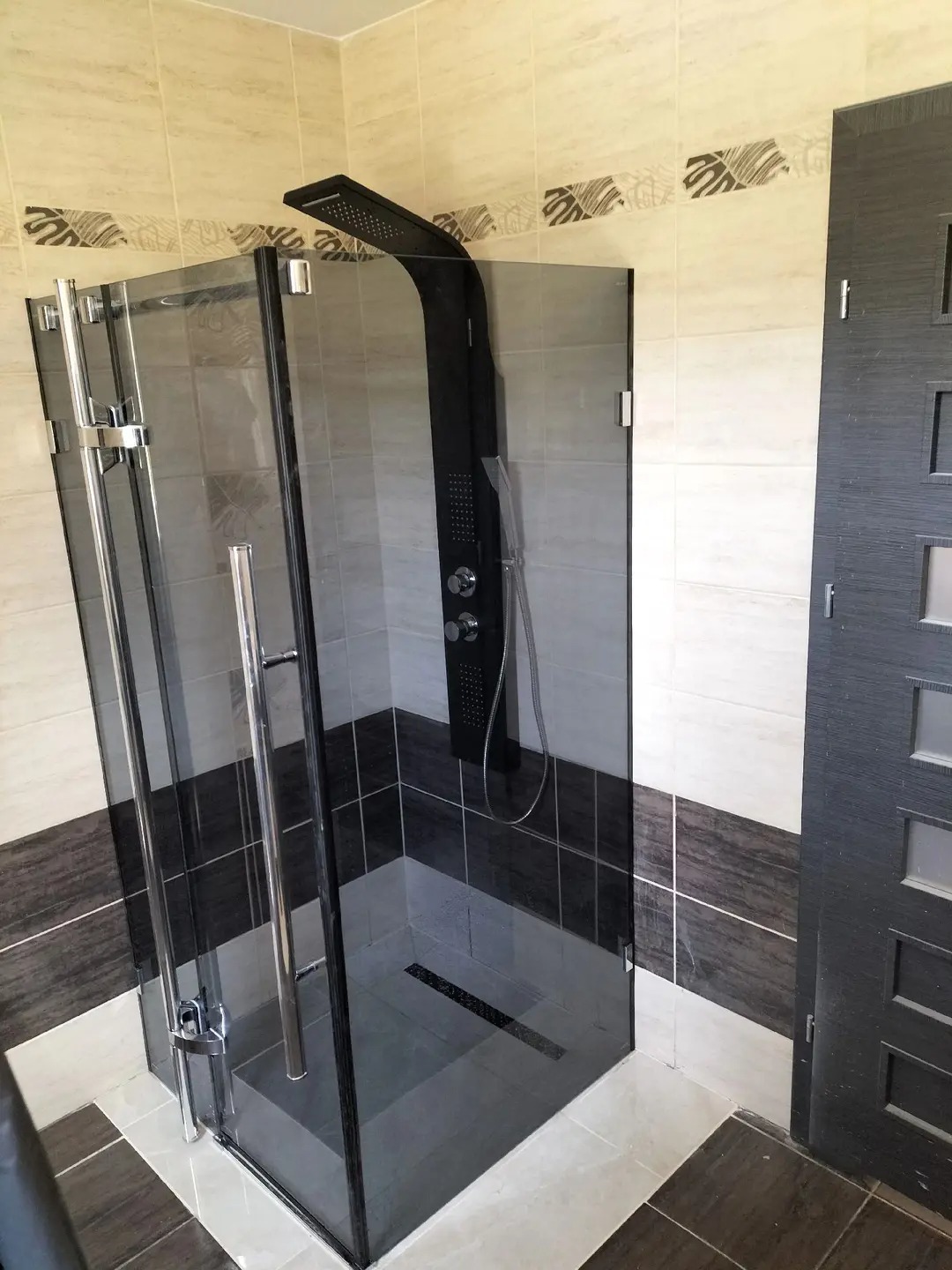 Nowoczesna kabina prysznicowa z czarnym panelem natryskowym i szklanymi drzwiami, w łazience wykończonej płytkami w odcieniach beżu i brązu z dekoracyjnym pasem.