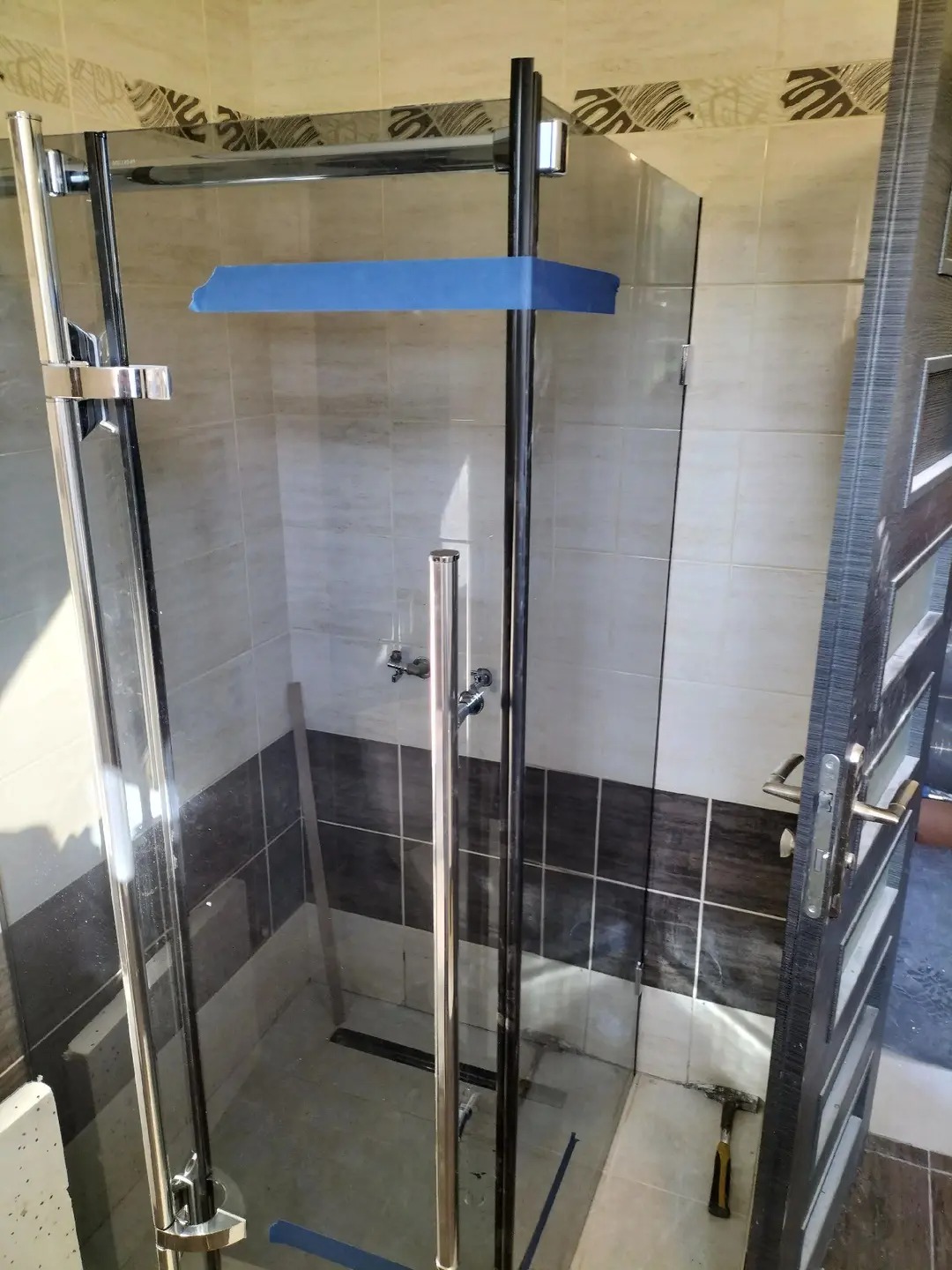 Montaż kabiny prysznicowej z chromowanymi profilami i szklanymi drzwiami, zabezpieczonymi niebieską taśmą. Widoczne płytki w odcieniach beżu i brązu oraz częściowo otwarte drzwi.