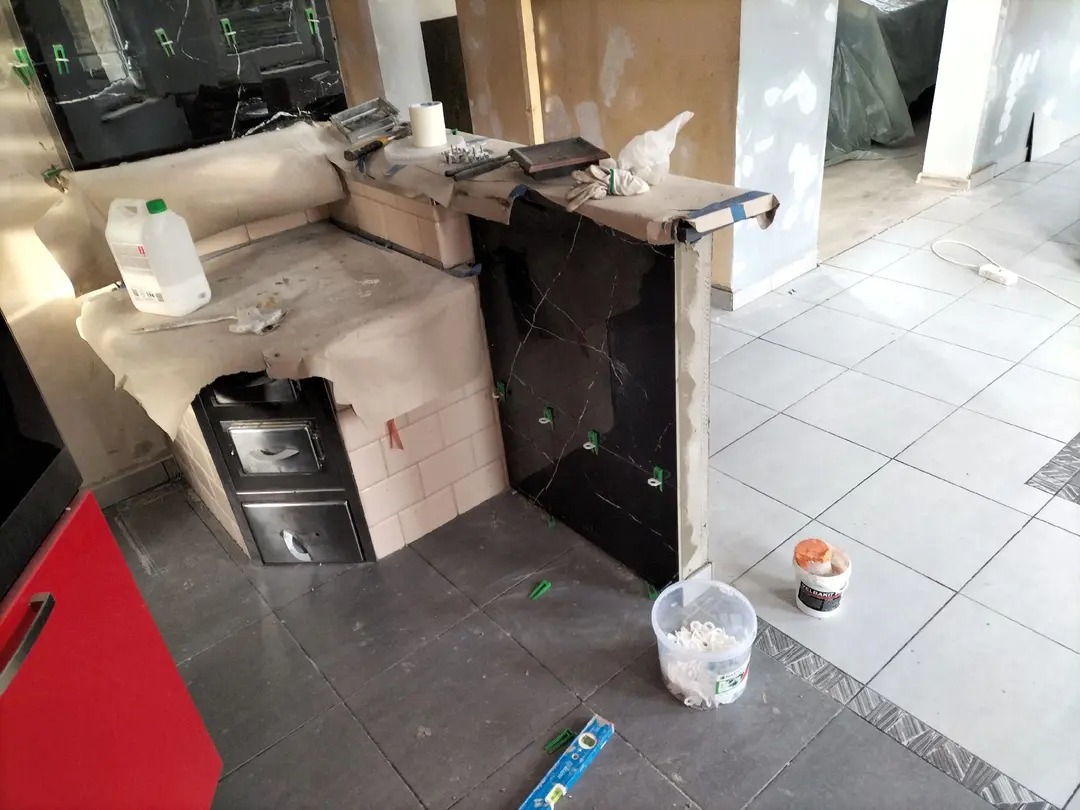 Układanie czarnej glazury imitującej marmur na ścianie i blacie w kuchni. Widoczne narzędzia i materiały glazurnicze, płytki podłogowe. Prace wykończeniowe w toku.