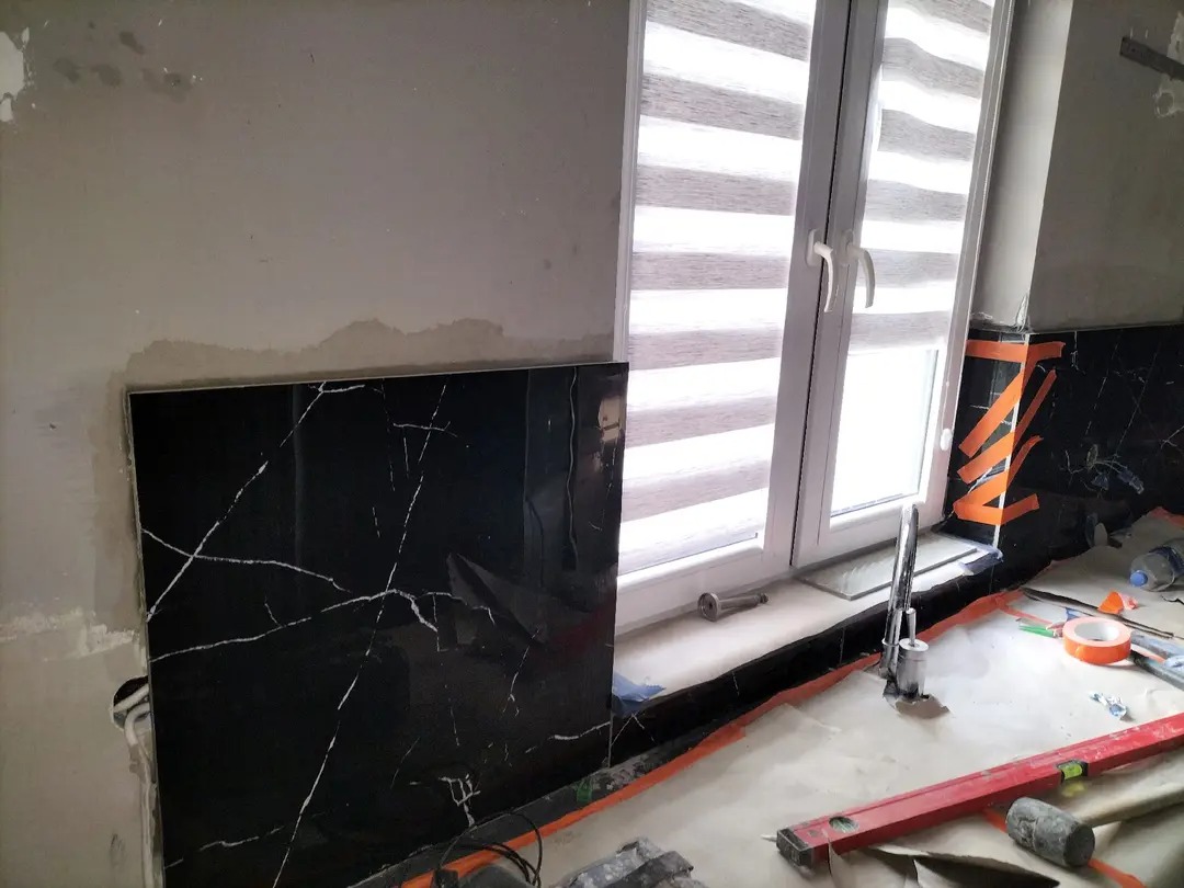 Montaż czarnej, marmurkowej płytki ściennej w łazience, widoczne narzędzia i okno z żaluzjami. Etap prac wykończeniowych.