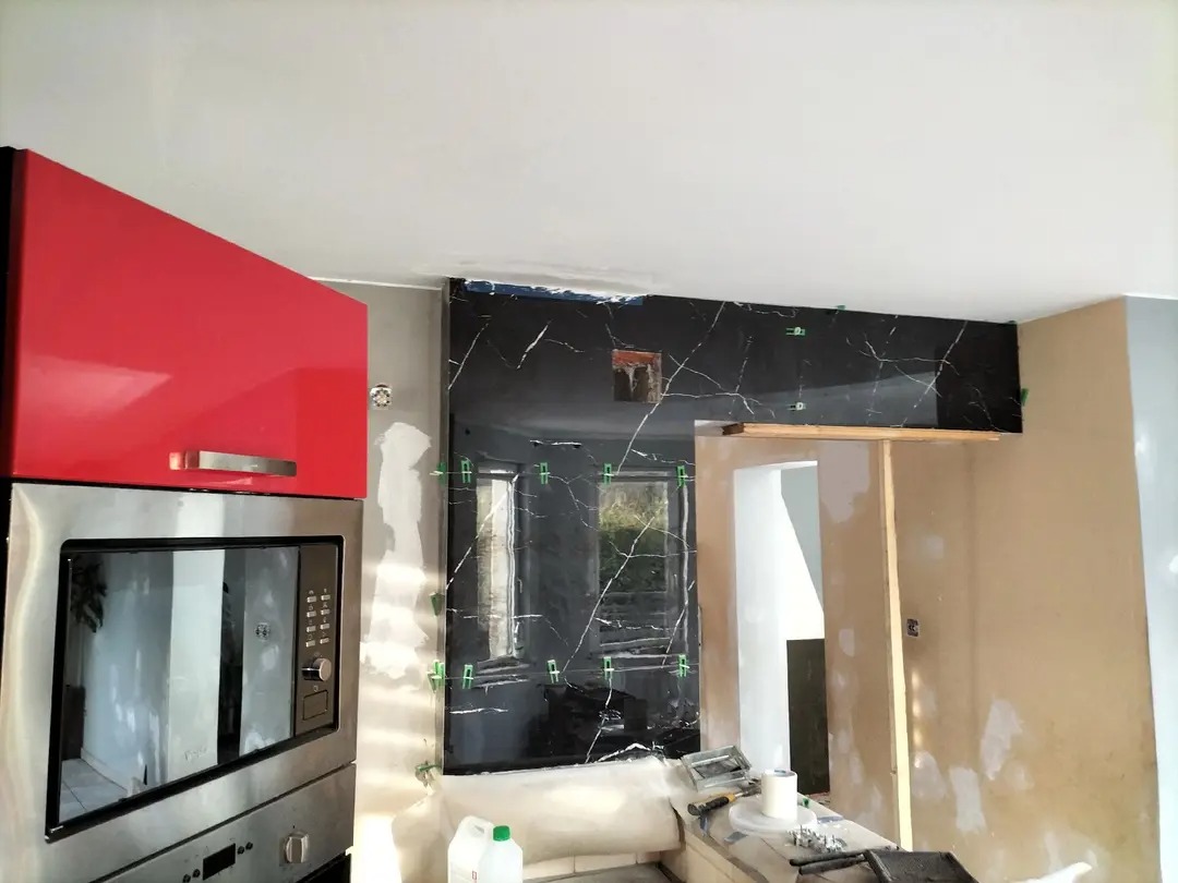 Częściowo wyłożona ściana czarnym marmurem z białymi żyłami w kuchni. Widoczne krzyżyki dystansowe, szafka i sprzęt AGD. Etap prac wykończeniowych.