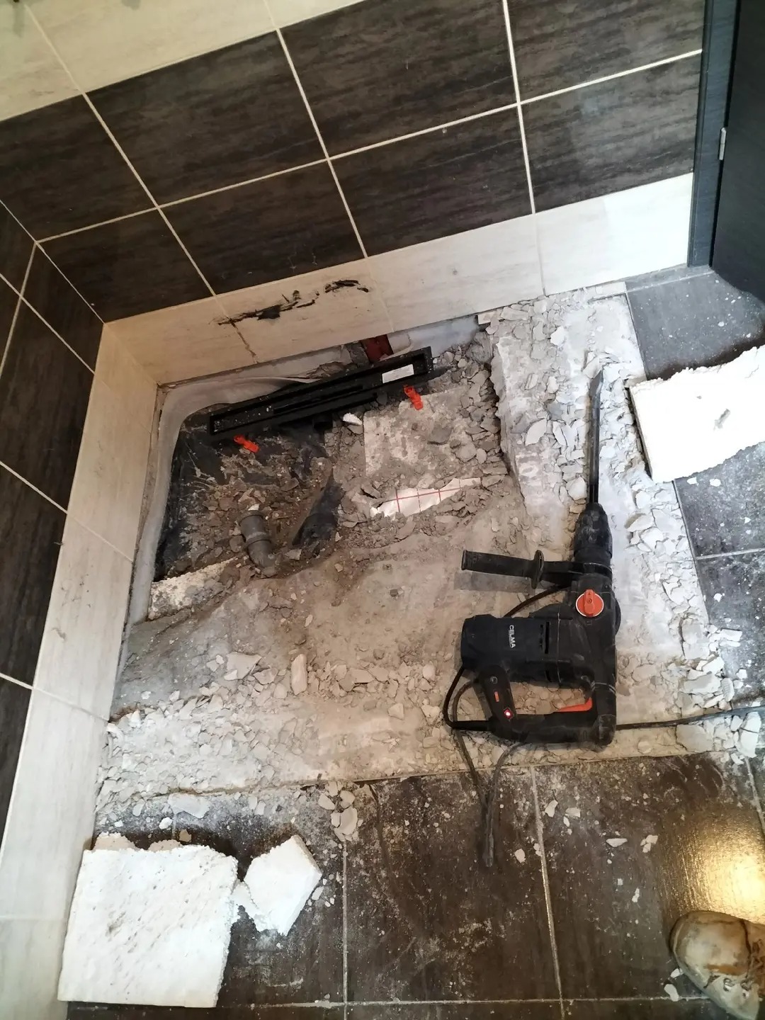 Demontaż starej posadzki w łazience z użyciem młota udarowego. Widoczne fragmenty płytek, gruz i instalacja odpływowa. Przygotowanie do remontu.
