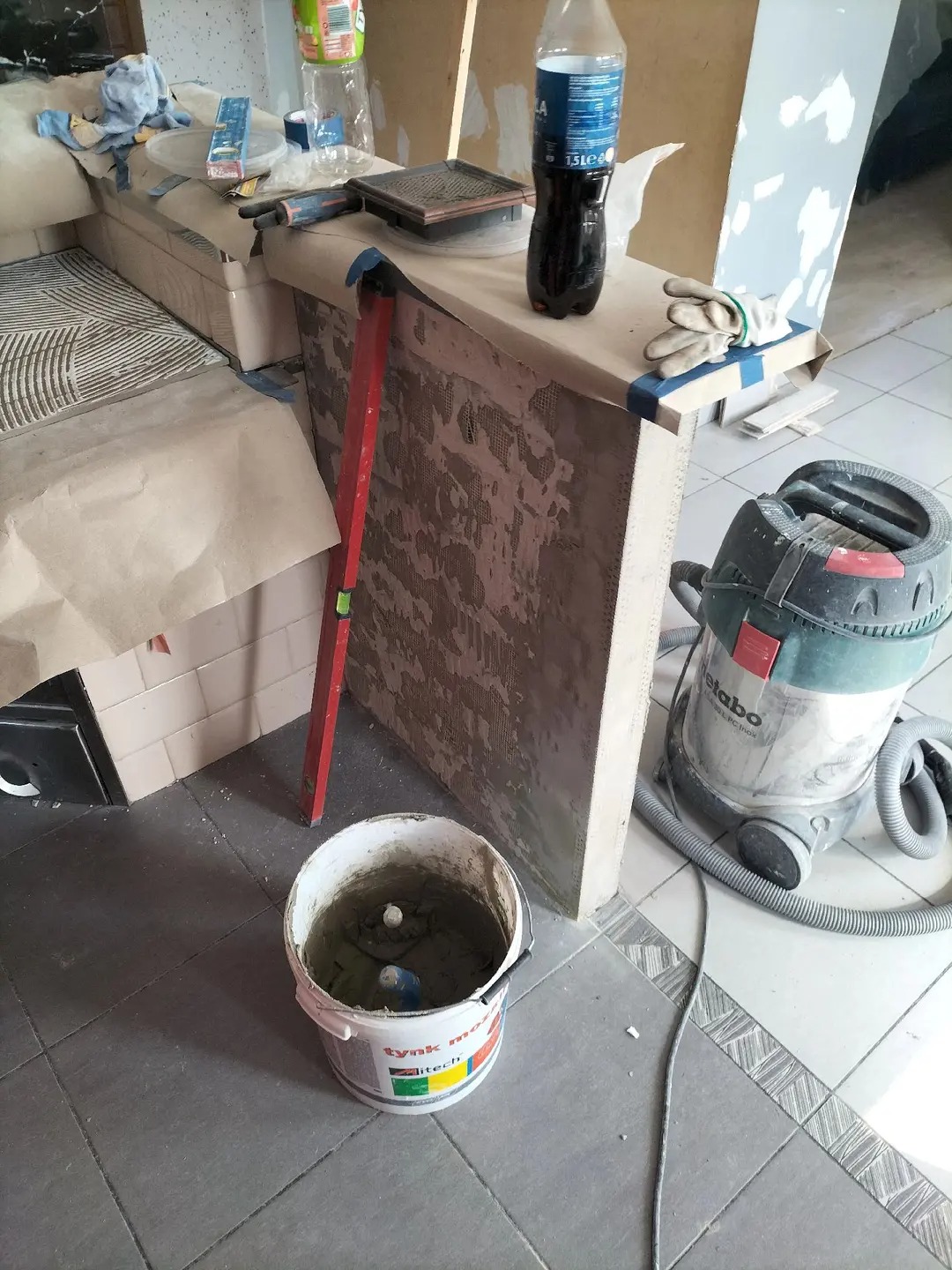 Wnętrze w trakcie prac glazurniczych: widoczny fragment ściany z przygotowaną powierzchnią pod płytki, poziomica, wiadro z zaprawą i odkurzacz przemysłowy.