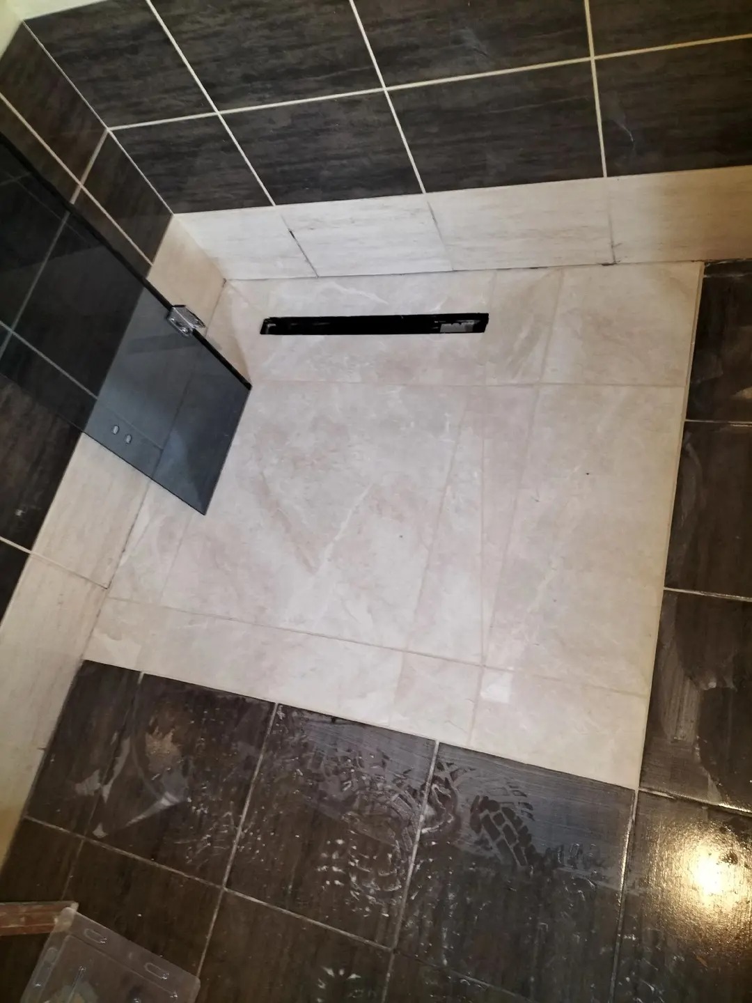 Ułożone jasne płytki podłogowe w łazience z odpływem liniowym, otoczone ciemnymi płytkami na ścianach i podłodze. Widok z góry, ukośne ujęcie.