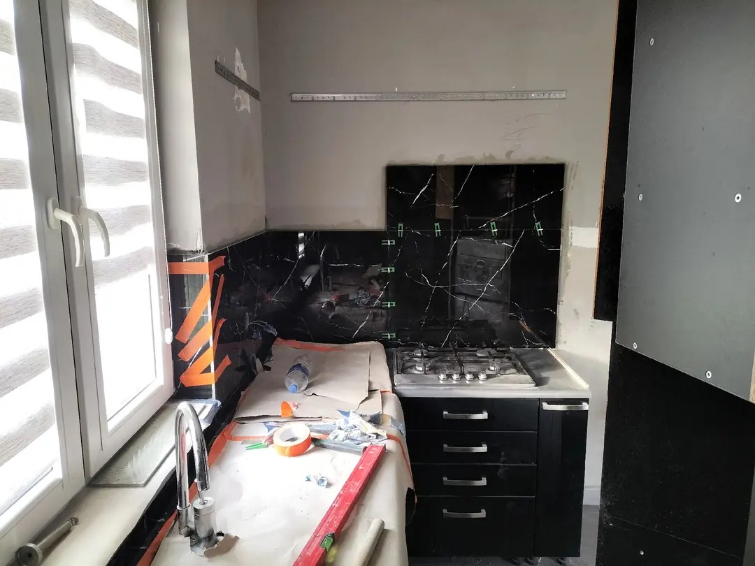 Fragment kuchni w trakcie montażu czarnych płytek imitujących marmur, widoczne narzędzia i poziomica, okno z żaluzjami.