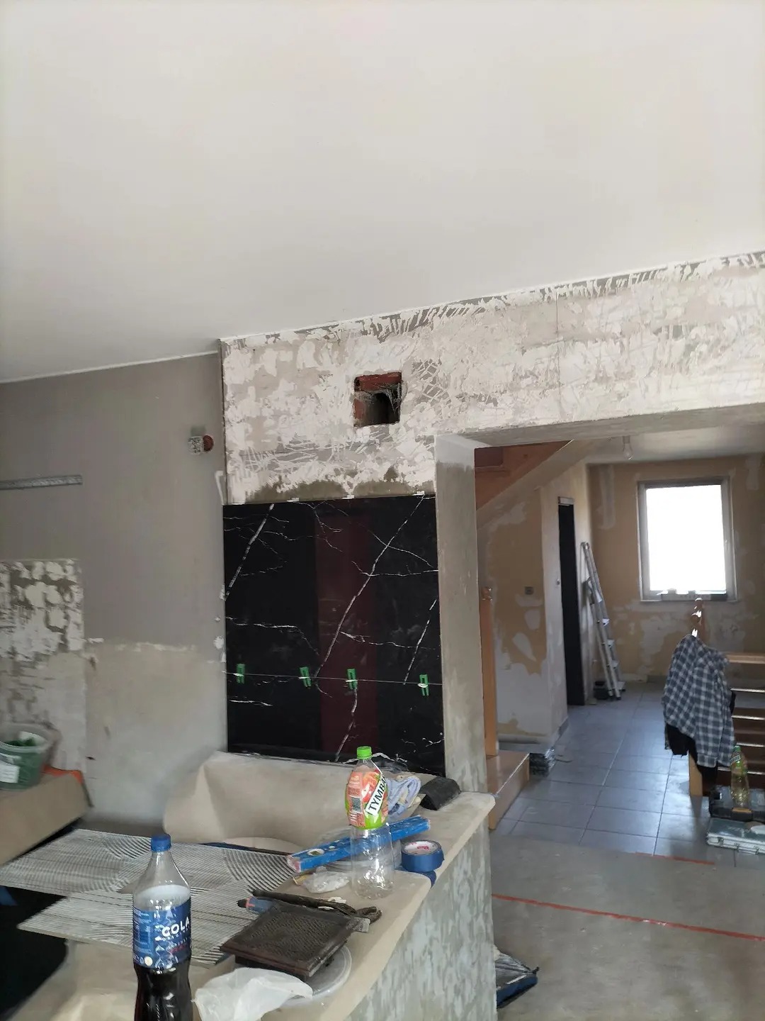 Częściowo położone, czarne płytki marmurowe na ścianie, zabezpieczone klamerkami. W tle widoczny remontowany pokój z drabiną i odkrytą cegłą.