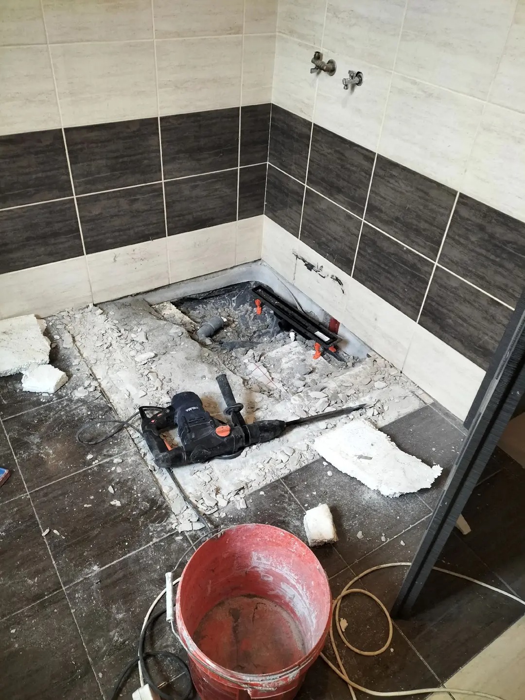 Demontaż płytek w łazience: widoczny gruz, narzędzia (młot udarowy), czerwone wiadro. Ściany z dwukolorową glazurą. Etap przygotowawczy do remontu.