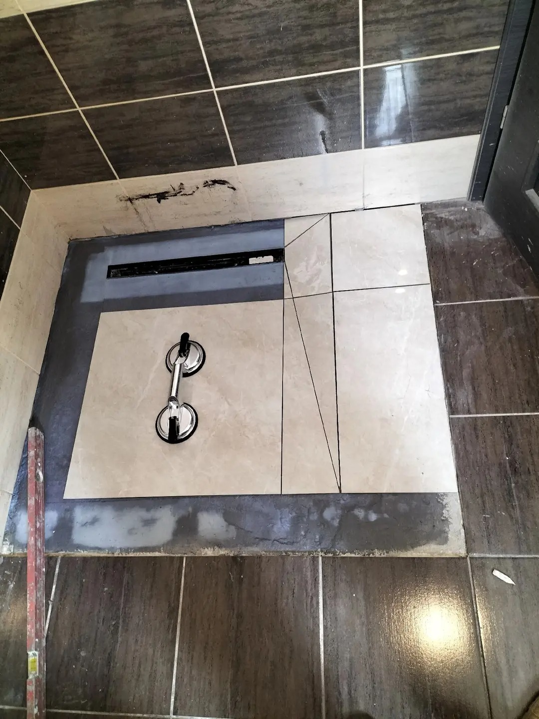 Montaż płytek w kabinie prysznicowej z odpływem liniowym. Widoczne narzędzie do przenoszenia płytek i poziomica przy ścianie. Górne płytki w odcieniach szarości.