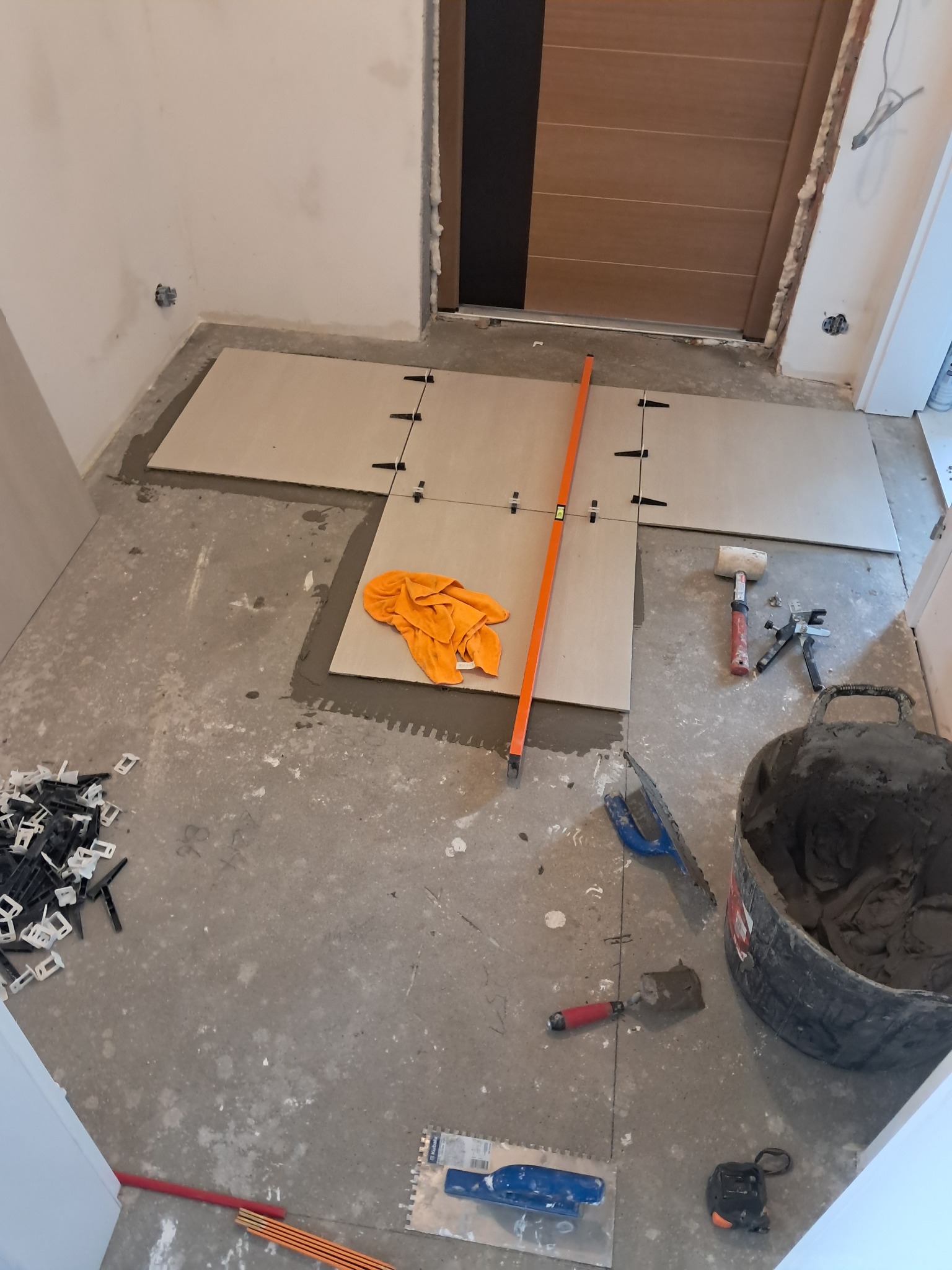 Układanie jasnych płytek podłogowych z użyciem poziomicy i krzyżyków dystansowych. Widoczny kubeł z zaprawą, kielnia i gumowy młotek. Prace glazurnicze w trakcie.