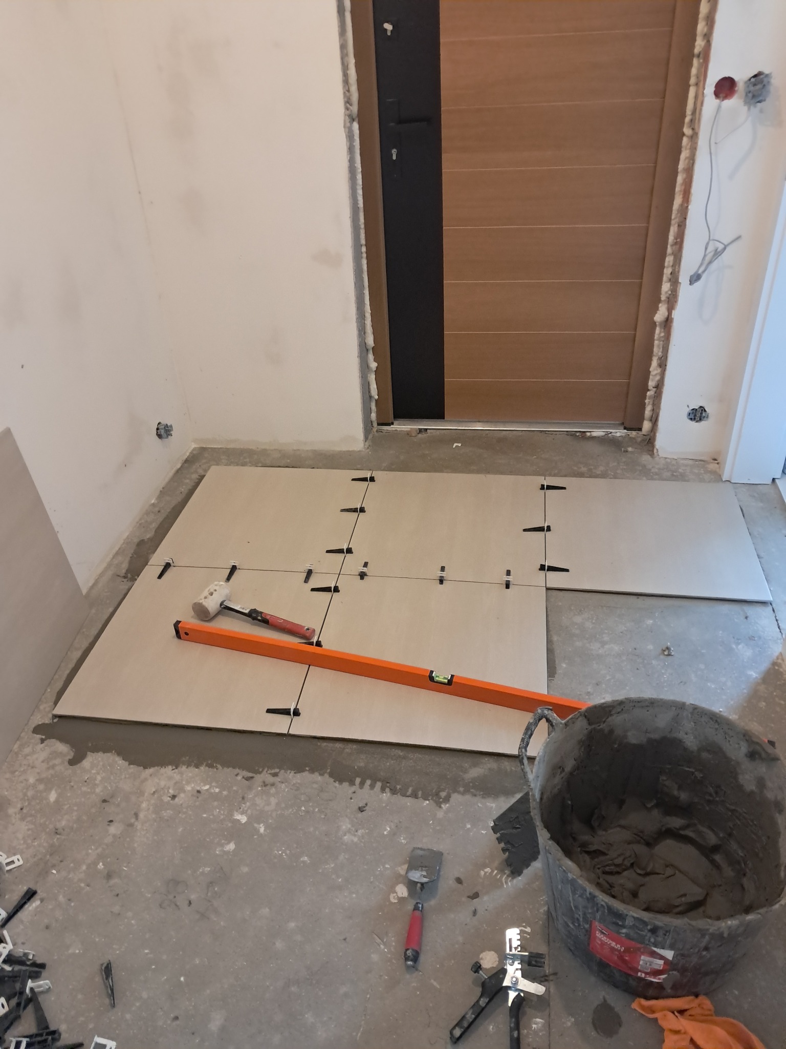 Układanie jasnych płytek podłogowych w korytarzu, widoczne narzędzia: poziomica, młotek, wiadro z zaprawą. Etap prac przed fugowaniem, krzyżyki dystansowe.